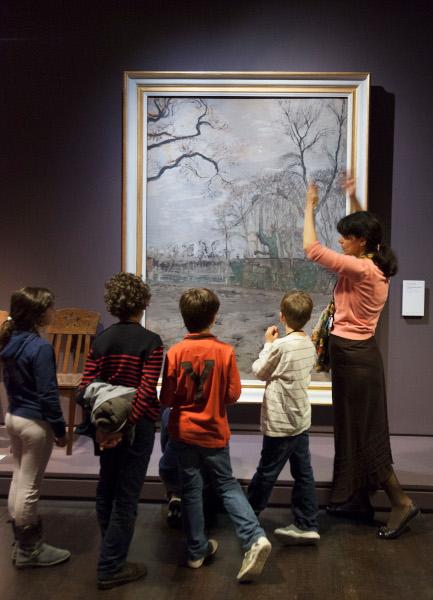Devant un tableau d'un paysage d'hiver, une femme brune décris l'oeuvre à quatre enfants
