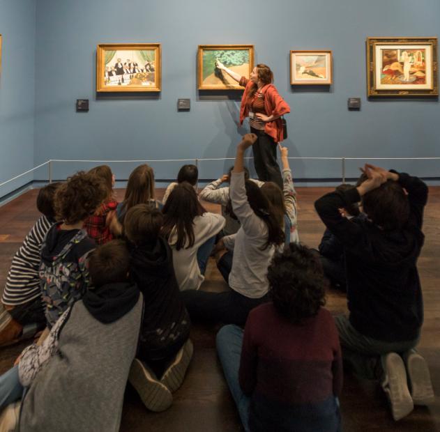 Devant des peintures impressionistes, une femme désigne et décris les détails d'une oeuvre à une dizaine d'enfants assis en tailleur sur le sol. 